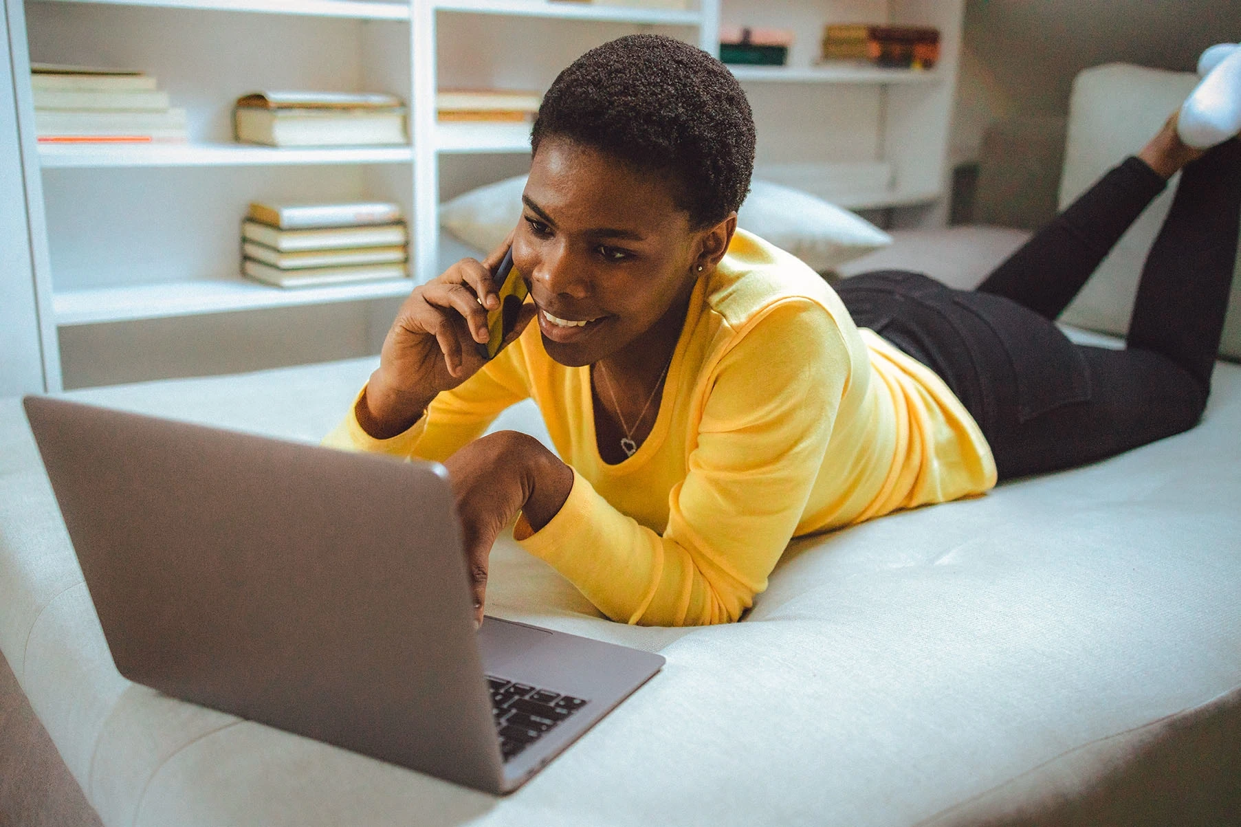 A young woman laying on her bed while on her phone, looking at a laptop.